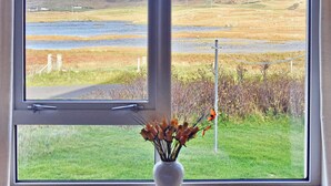 A window with a view of a grassy landscape, a body of water, and a hill. - 2 bedroom accommodation in Howbeg (Tobha Beag), Isle of South Uist (Howbeg (Tobha Beag), Isle of South Uist)