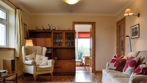 A cozy living room with a wooden cabinet, a beige armchair, a sofa with floral cushions, and a rug. - 2 bedroom accommodation in Howbeg (Tobha Beag), Isle of South Uist (Howbeg (Tobha Beag), Isle of South Uist)