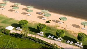 An aerial view of a beach with rows of umbrellas and lounge chairs, a pier, and a boat creating a wake in the water. - Salem Apartment (Ismailia)