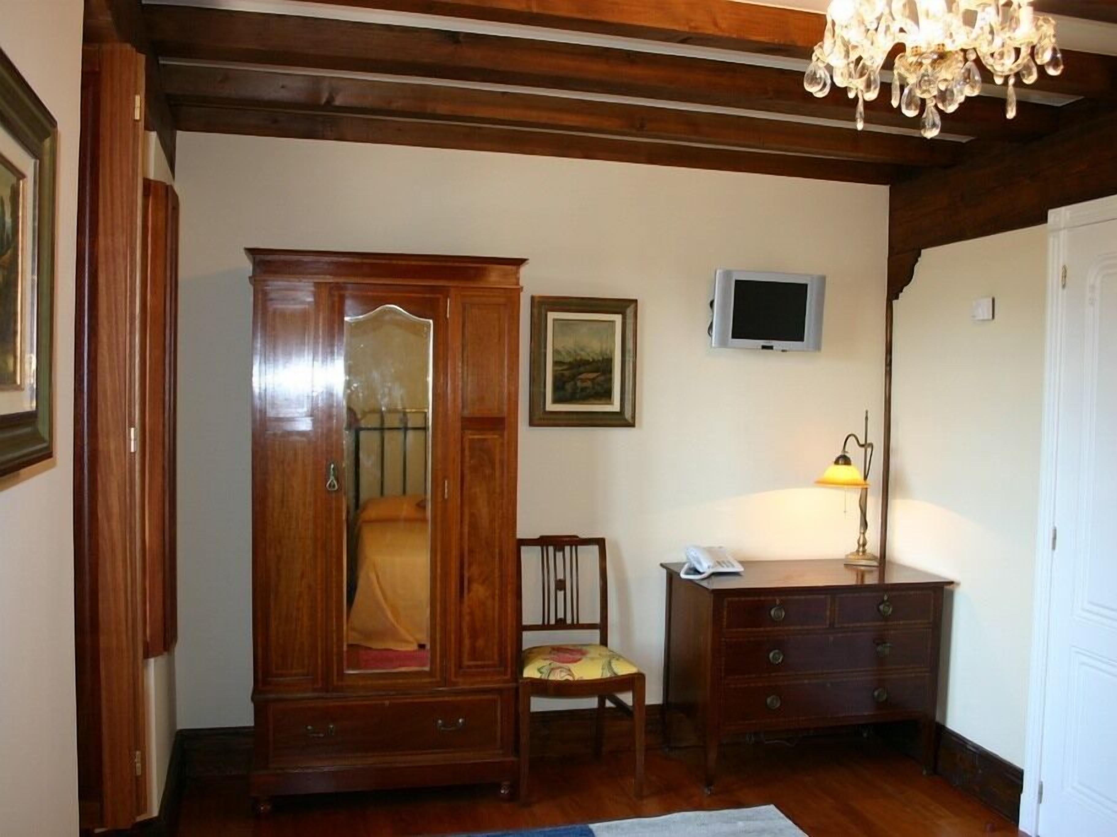 Quarto com guarda-roupa de madeira, uma cadeira de madeira pequena, uma escrivaninha com luminária e uma televisão na parede.