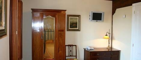 Quarto com guarda-roupa de madeira, uma cadeira de madeira pequena, uma escrivaninha com luminária e uma televisão na parede.