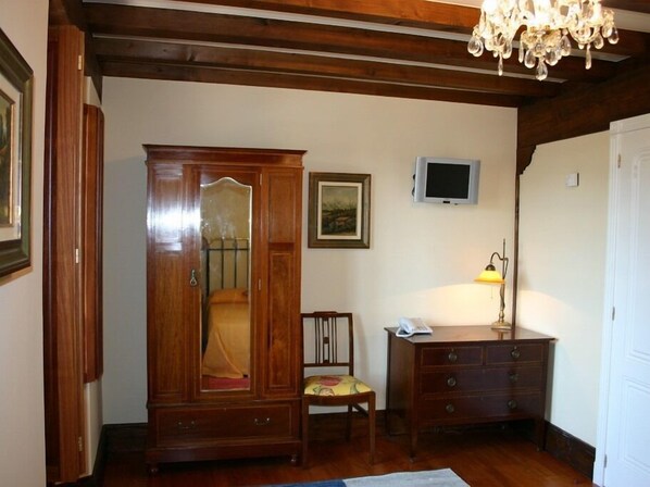 A room with a wooden wardrobe, a small wooden chair, a desk with a lamp, and a television on the wall. - Posada el Hidalgo Mingucho (Medio Cudeyo)