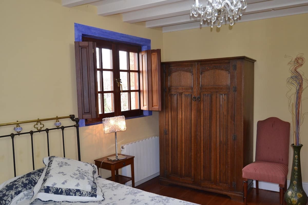 A bedroom with a bed, a wooden wardrobe, a chair, a lamp, and a window with shutters.