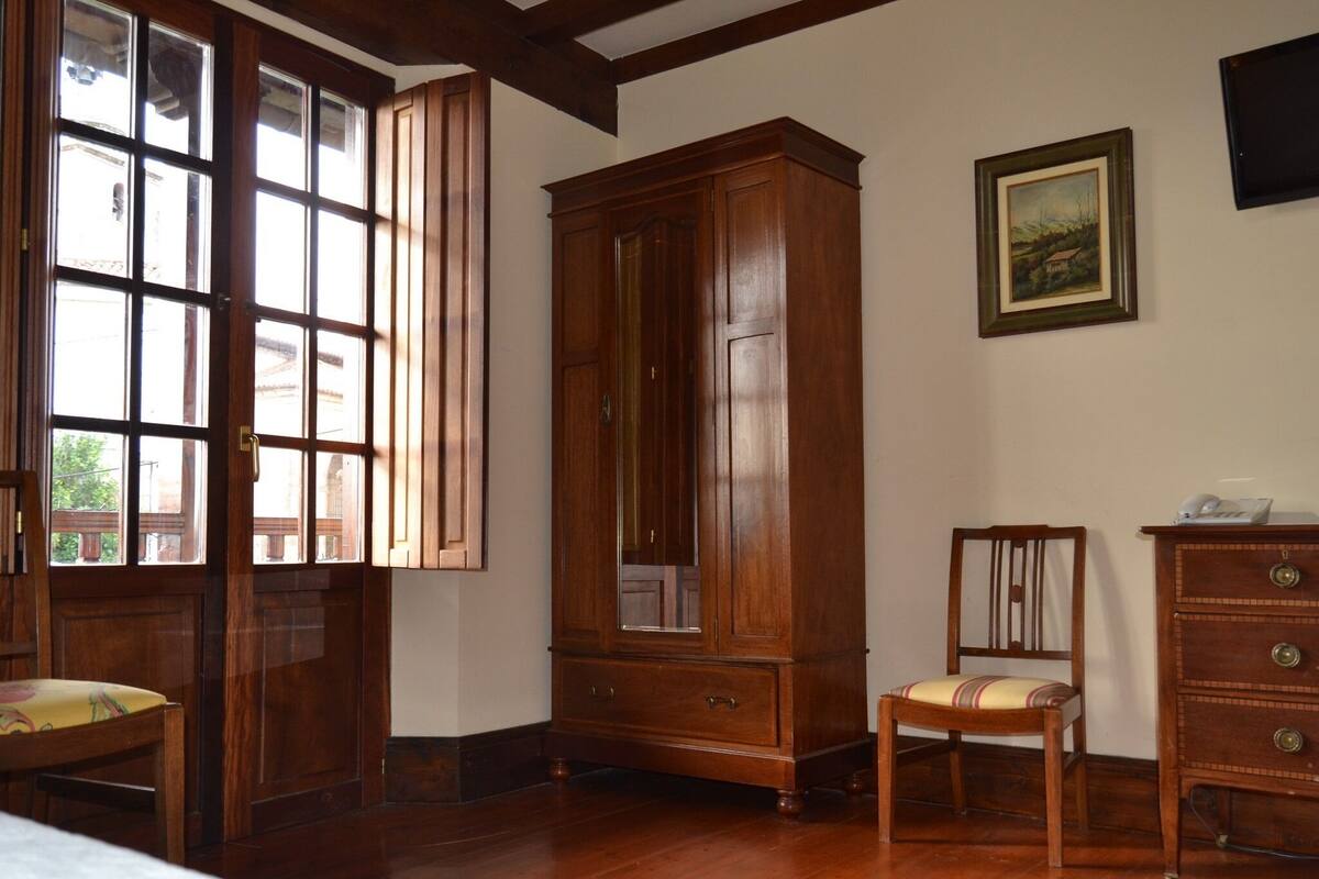 A room with wooden flooring, a large wooden cabinet, a chair, a small table, and a painting on the wall.