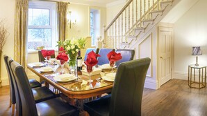 A dining room with a wooden table set for four, surrounded by chairs, a chandelier, and a staircase in the background.