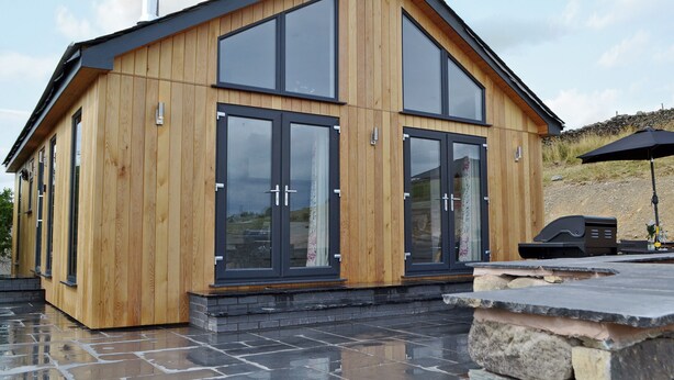 Une cabane moderne en bois, dotée de grandes portes vitrées et d’une terrasse en pierre.