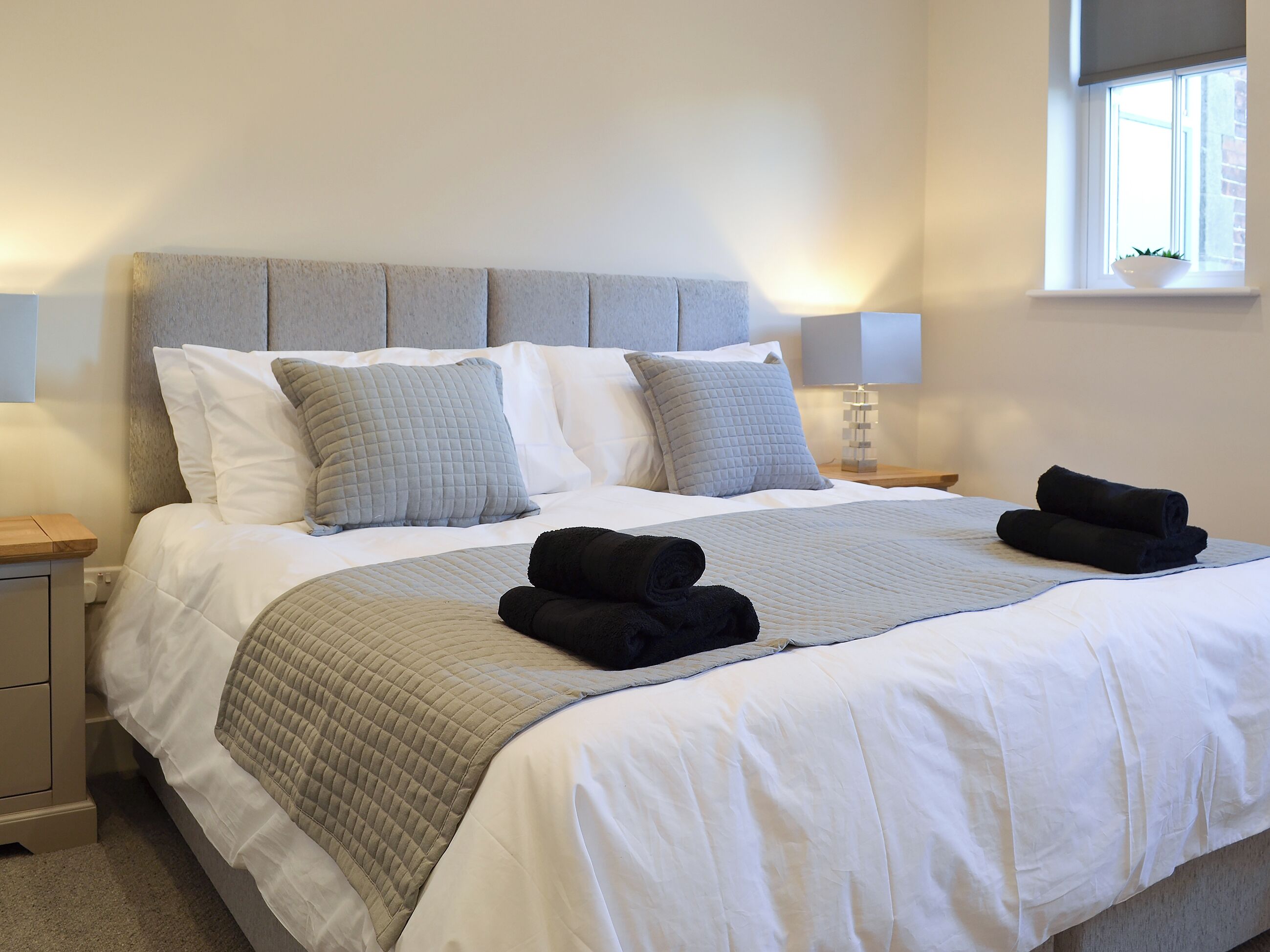 A neatly made bed with a grey headboard, white bedding, and two black towels on the side.