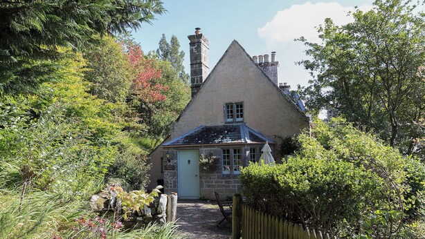 A stone house with a steeply pitched roof, a visible chimney, and a small porch area.