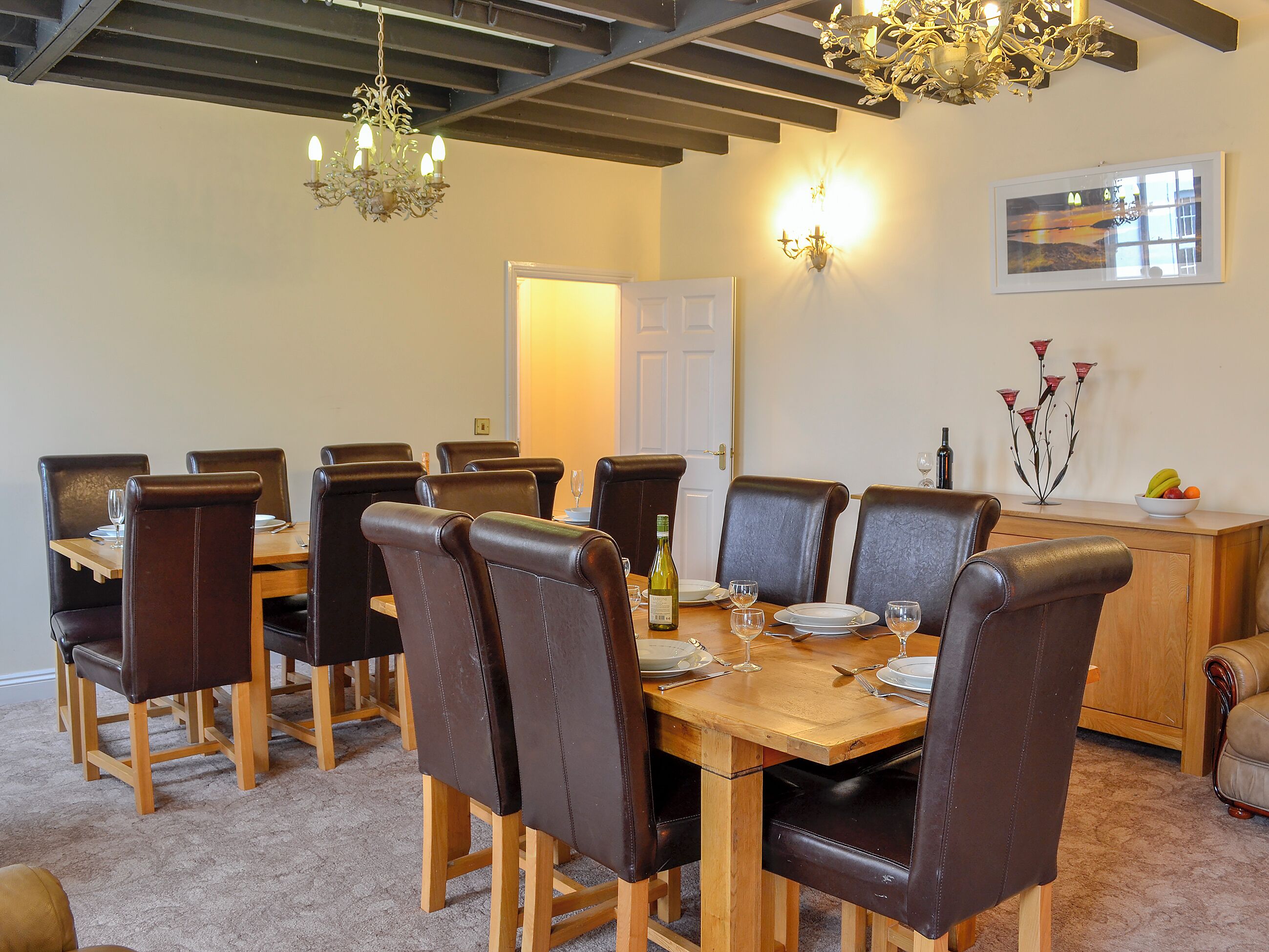 A dining area with a wooden table and leather chairs, a chandelier, and a fruit bowl on a sideboard.