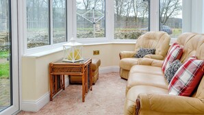 A conservatory with a beige leather sofa, a small table with a lantern, and a view of a garden through large windows. - Maesbangor Farmhouse (Capel Bangor, near Aberystwyth)