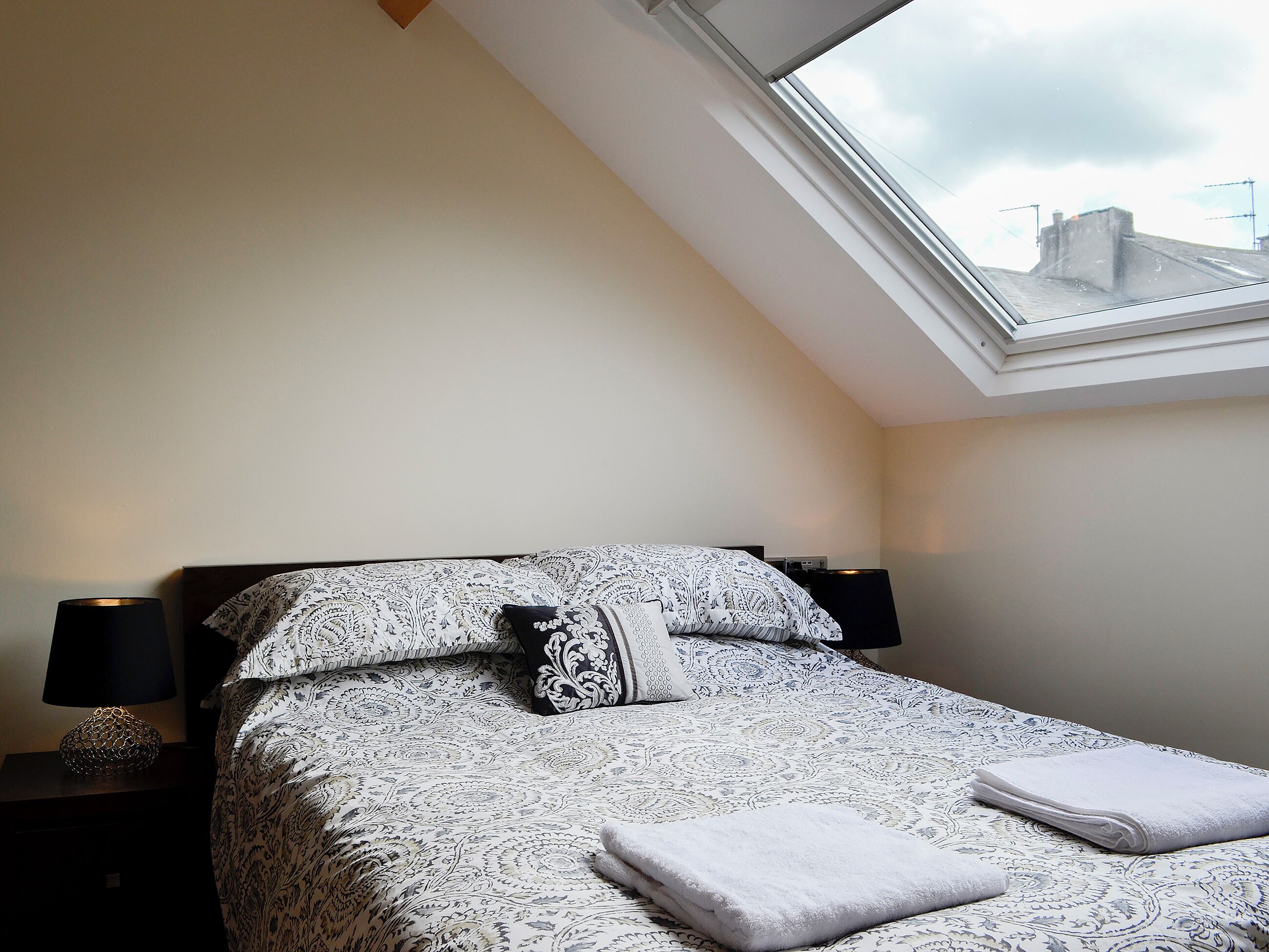 A bedroom with a skylight, a patterned bedspread, two pillows, a bedside lamp, and a folded towel on the bed.