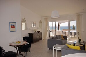 A modern living room with a sofa, a dining table, and a view of the sea through large windows.