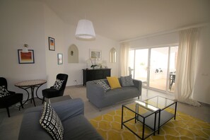 Uma sala de estar com sofá, poltronas, mesa de centro de vidro, uma mesinha e uma janela grande com cortinas.
