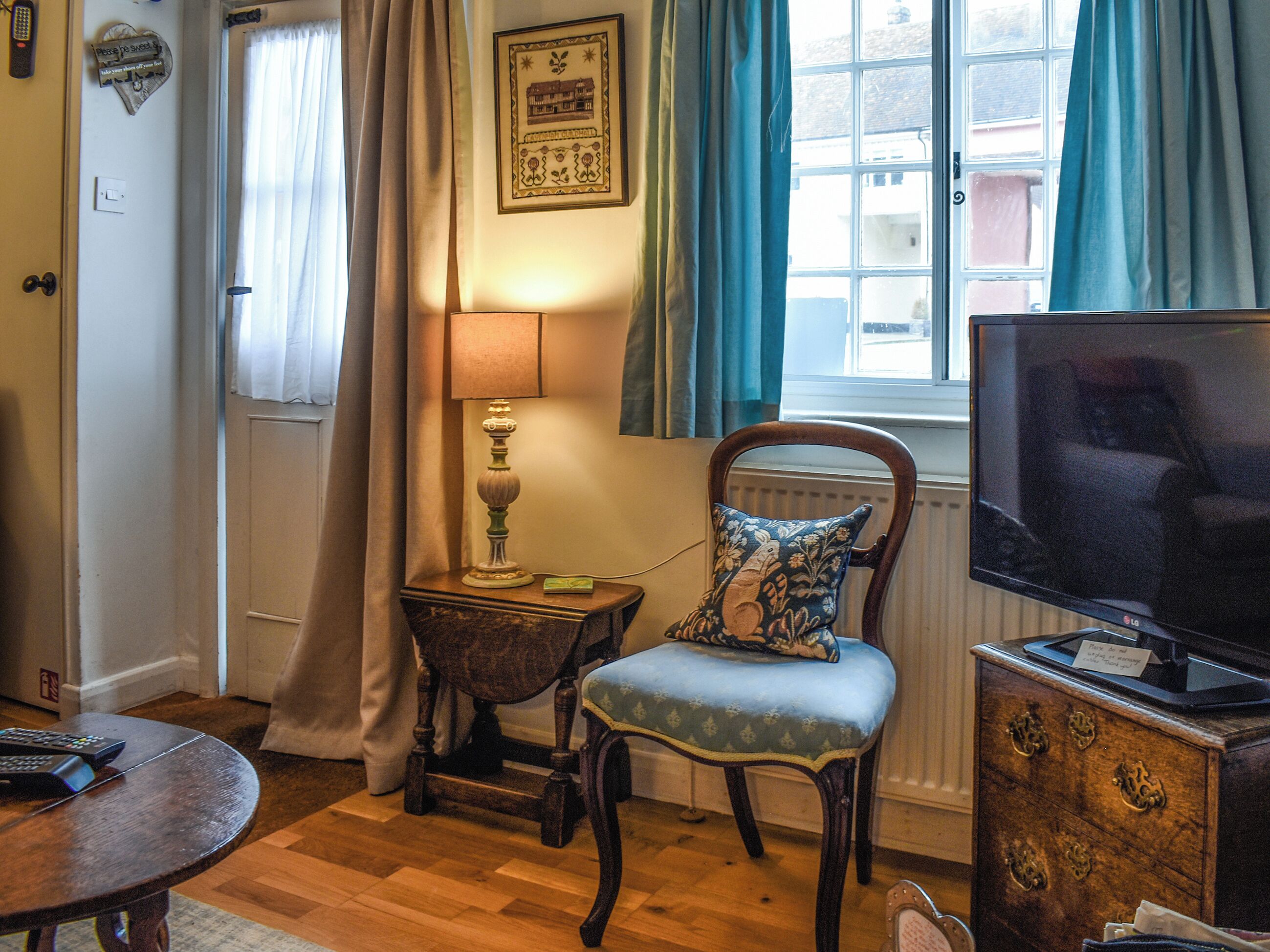 A cozy living room with a wooden floor, a television, a chair with a floral cushion, a small table with a lamp, and a window with curtains.