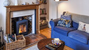 A cozy living room with a fireplace, a blue sofa, and a coffee table with remote controls. - Lavenham Red Brick Cottage (Lavenham)