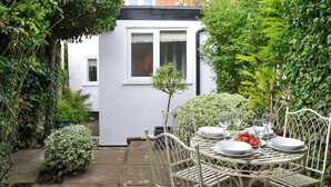A white metal table set with plates, glasses, and strawberries in a garden with white walls and a small tree. - Lavenham Red Brick Cottage (Lavenham)