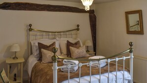A neatly made bed with white linens and brown throw pillows, a bedside table with a lamp, and a wooden beam above the bed. - Lavenham Red Brick Cottage (Lavenham)