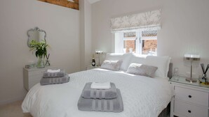 Un dormitorio con una cama grande, paredes blancas, una viga de techo de madera, una ventana con vista a un muro de ladrillo y una mesita de noche con una lámpara y una botella.