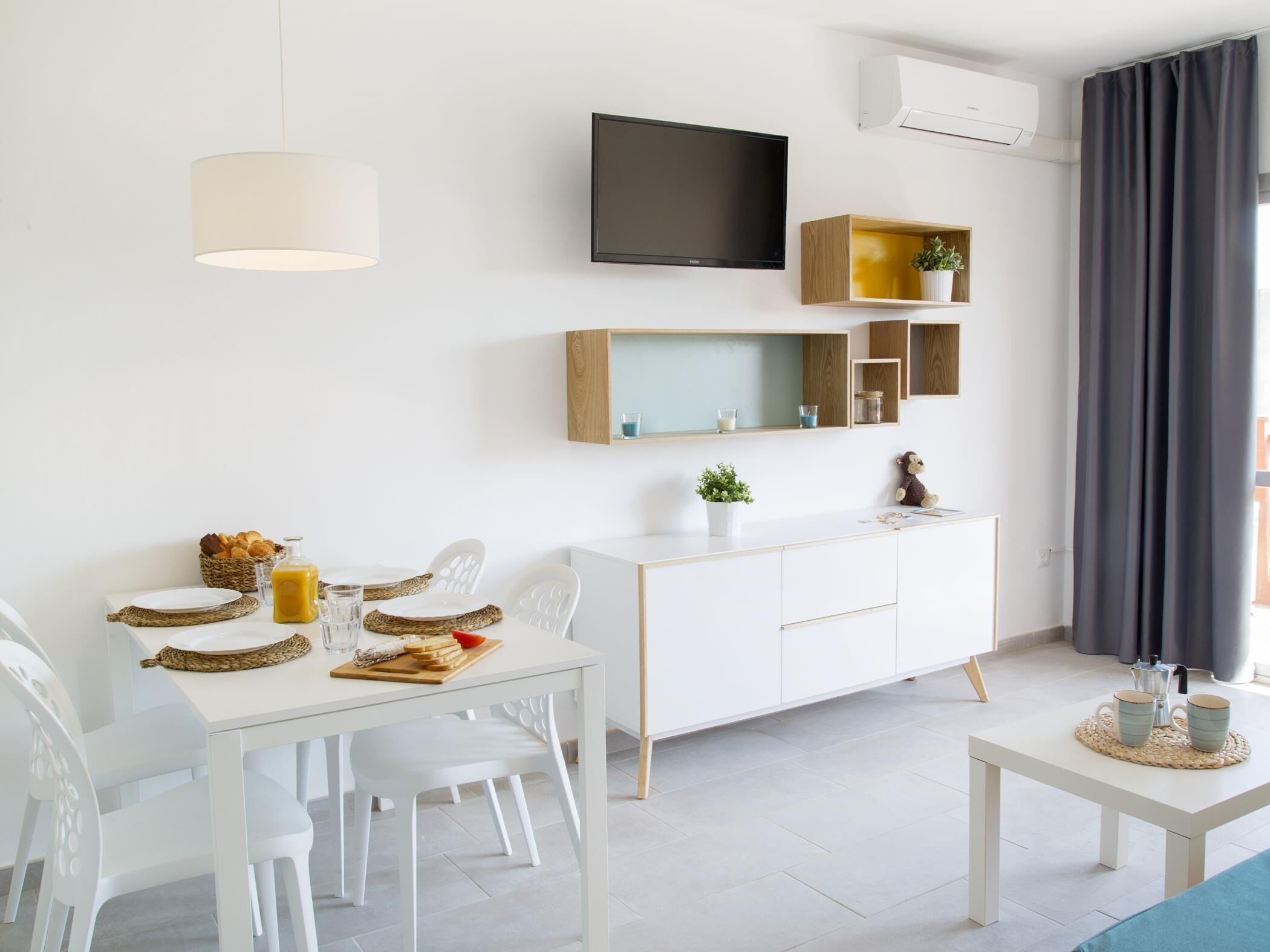 Un salon moderne et minimaliste avec une table à manger blanche, des chaises blanches, un meuble blanc, un téléviseur fixé au mur et une petite table d’appoint avec une plante et une tasse.