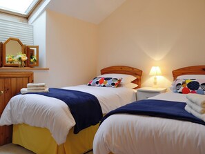A hotel room with two beds, each with a wooden headboard, white bedding, and a blue blanket. There is a nightstand with a lamp between the beds, and a wooden wardrobe with a mirror above it.