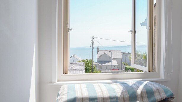 Une fenêtre donnant sur une ville côtière, un mur blanc et un coussin rayé posé sur une étagère en bois.