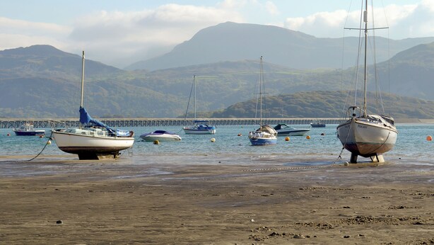 A beach with boats anchored near a pier, surrounded by hills.