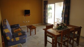 Ein Wohnzimmer mit einer Couch, einem Esstisch mit Stühlen, einem an der Wand montierten Fernseher und Blick ins Freie durch eine Schiebetür.