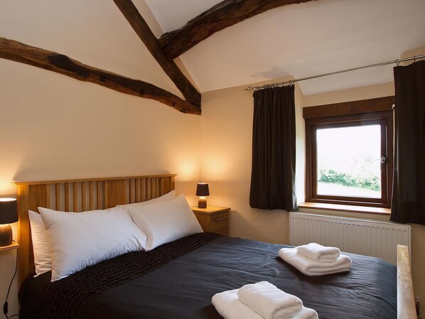 A bedroom with a wooden headboard, a bed with white and dark bedding, a nightstand with a lamp, a window with curtains, and a radiator. - Damson Cottage - LM1 (Waberthwaite, near Ravenglass)