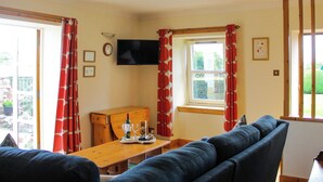 Ein Wohnzimmer mit einer Couch, einem Holztisch mit Flasche und Gläsern, einem Fernseher an der Wand und einem Fenster mit Vorhängen.