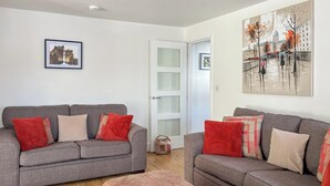 Ett vardagsrum med två grå soffor, röda och beige kuddar, ett trägolv, en rosa pall och en tavla som föreställer en stadsbild.