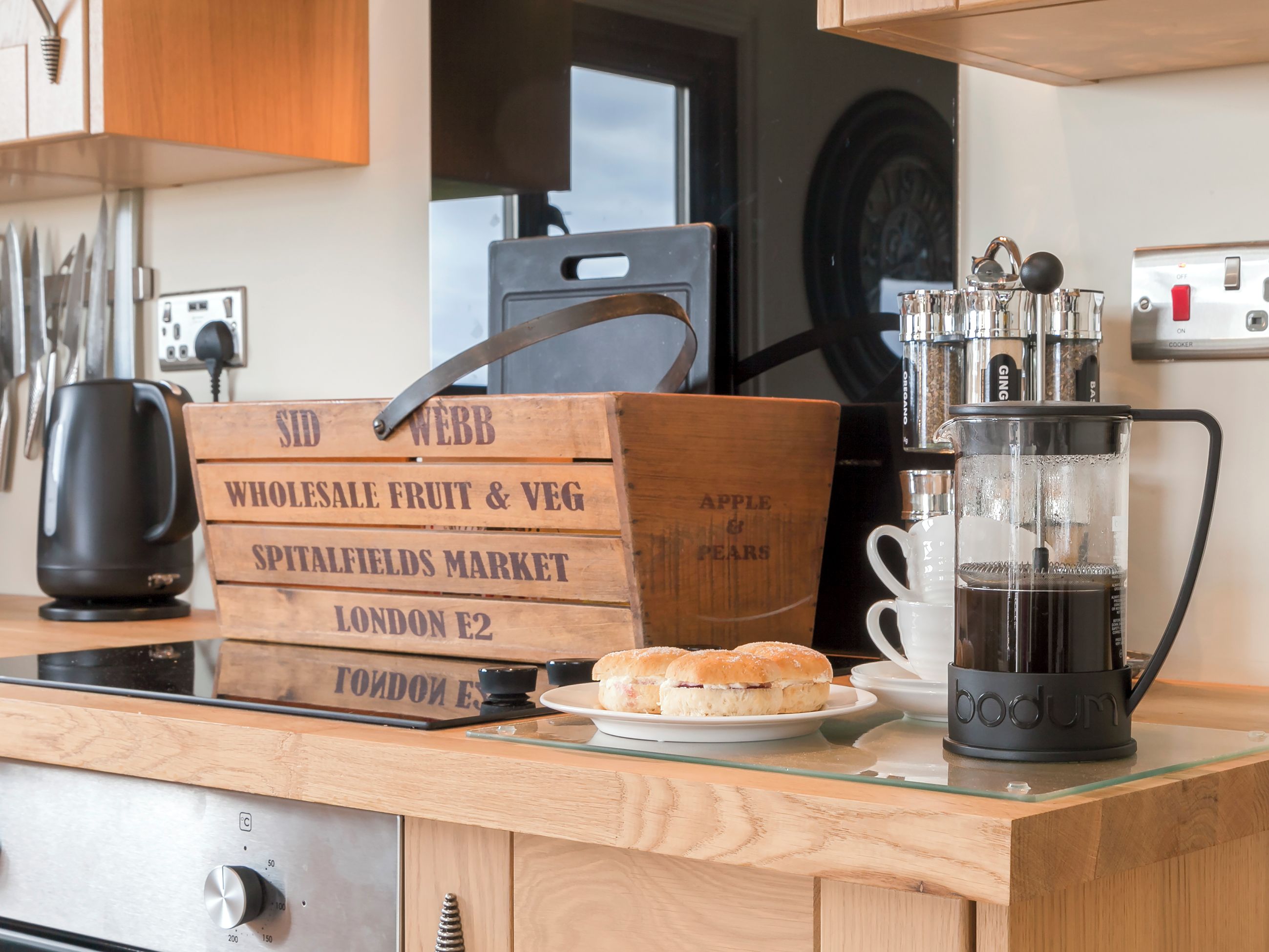 A kitchen counter with a coffee maker, a wooden crate labeled 'Whole Sale Fruit & Veg', and a plate of sandwiches.