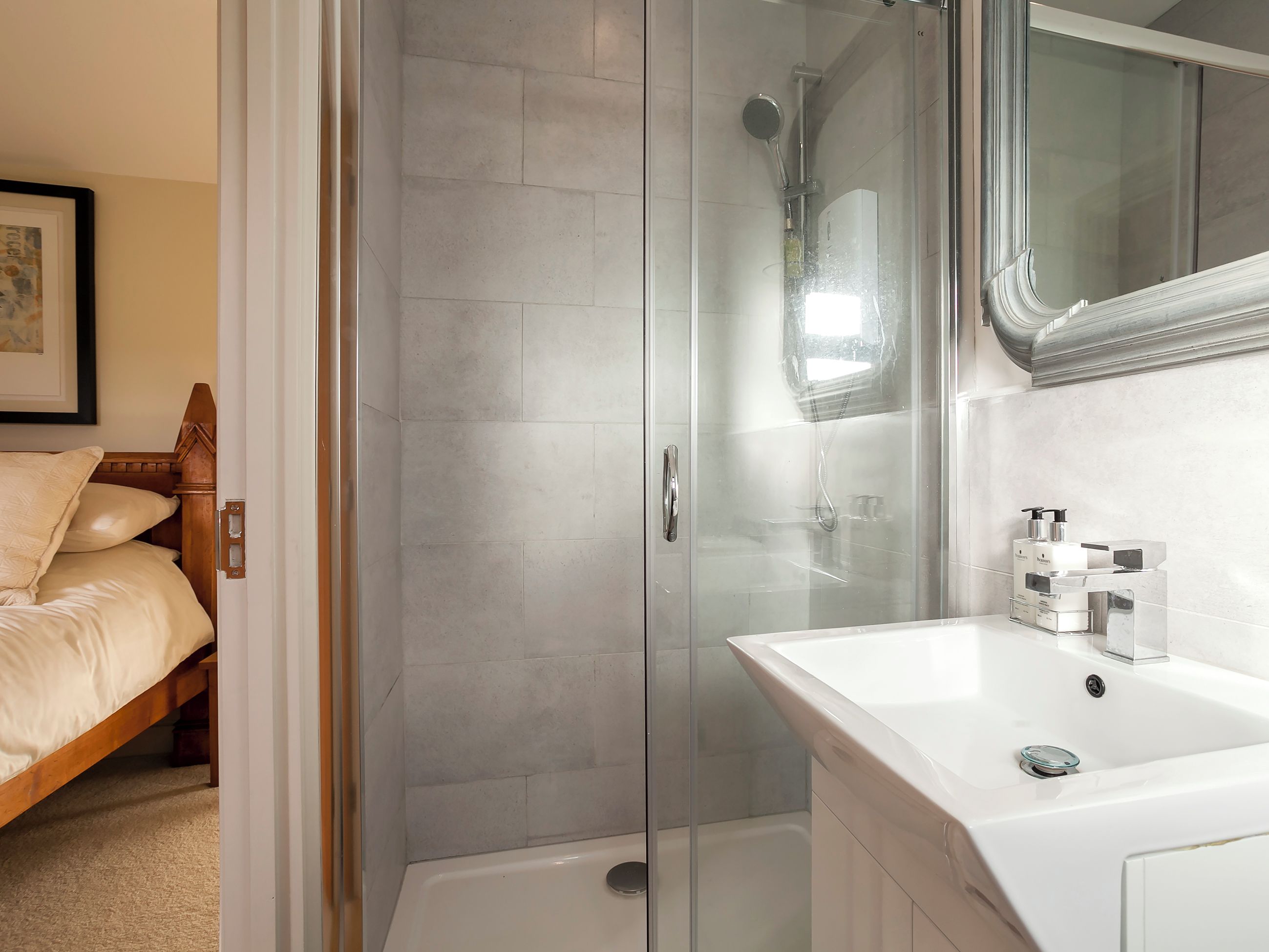 Un baño moderno con ducha envuelta en vidrio, un lavamanos blanco con espejo y una cama de madera en la habitación contigua.