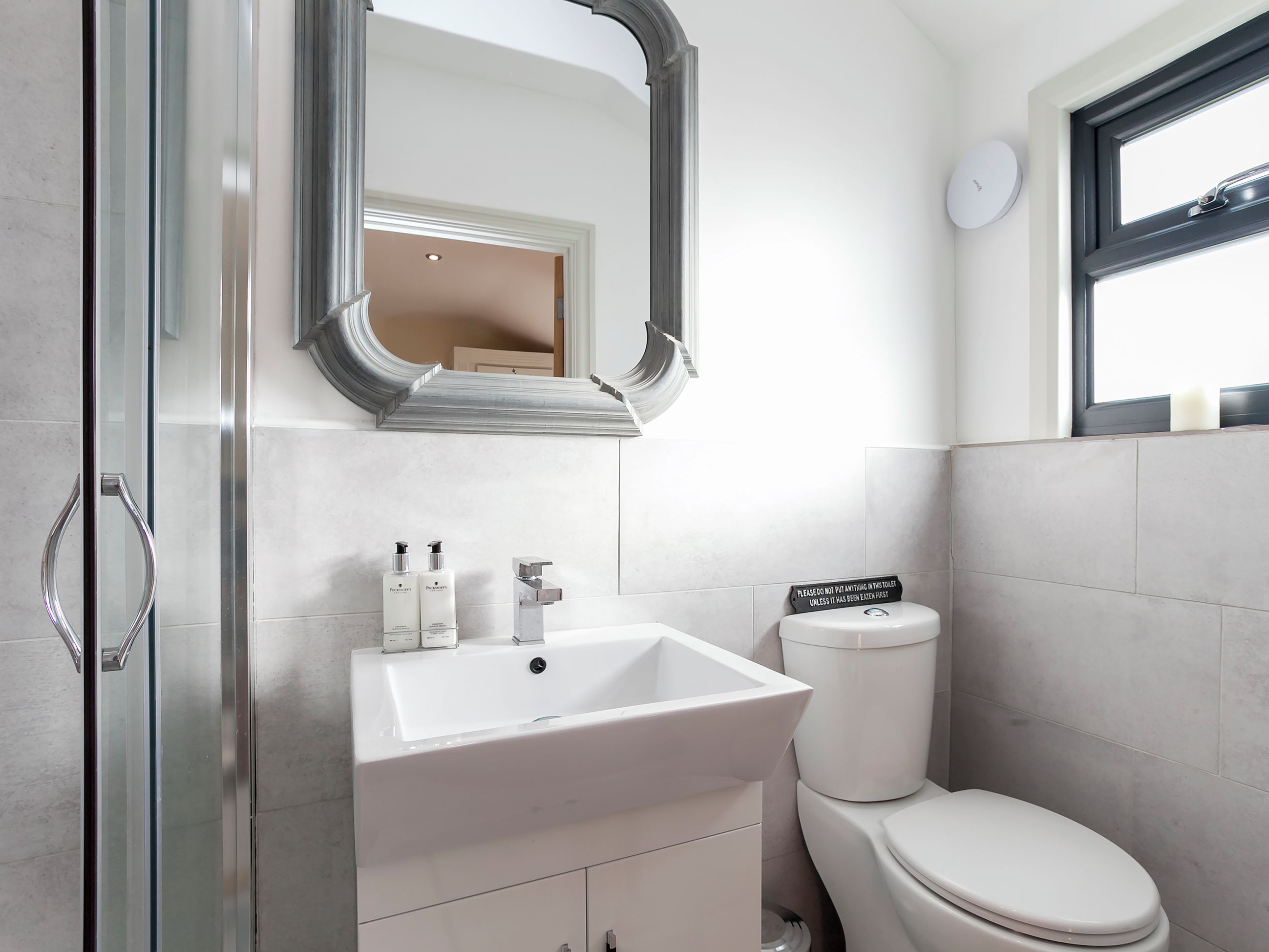 Un baño moderno con cabina de ducha de vidrio, un lavabo blanco con dos botellas de champú, un inodoro y un espejo con marco decorativo.