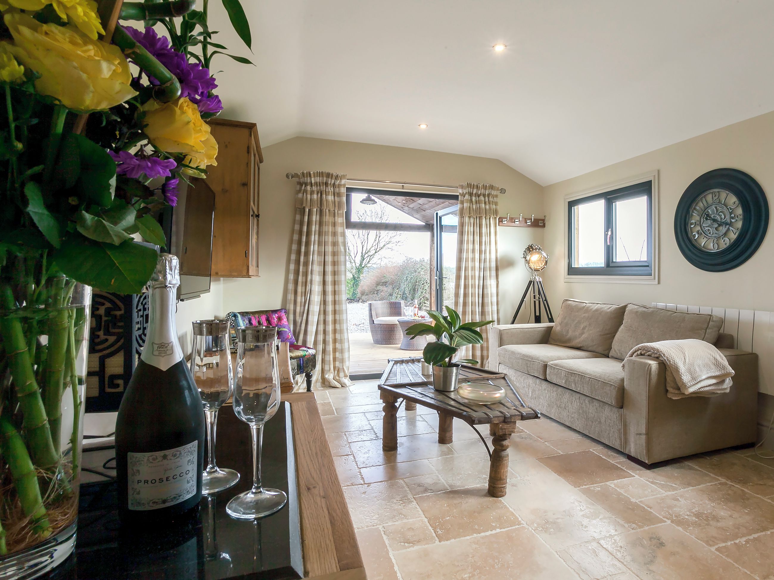 A living room with a sofa, coffee table, and a bottle of wine. There is a window with curtains and a view of the outside.