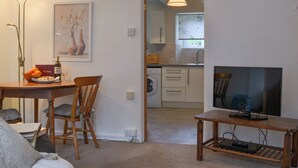 A living room with a TV, a dining area with a table and chairs, a kitchen in the background, and a hallway leading to another room. - 1 bedroom accommodation in Lostwithiel (Lostwithiel)