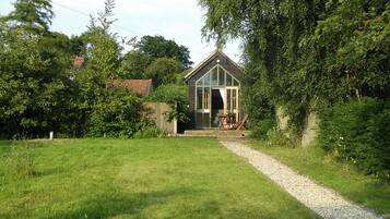 A small, rustic wooden structure with a sloped roof, surrounded by lush greenery and a gravel path leading to it.