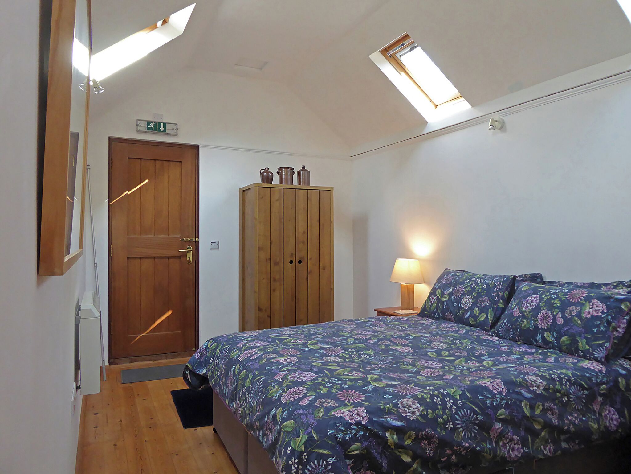 Une chambre avec une armoire en bois, un lit avec une literie à motifs floraux, une table de chevet avec une lampe et des fenêtres de toit.