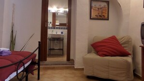 A room with a bed, a chair, a small table, a mirror, and a bathroom visible through an archway. - Historic house 3 bedrooms 3 bathrooms with panoramic terrace (CASTELSARDO)