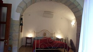 A bedroom with a stone archway, a bed with a red bedspread, a nightstand with a lamp, and a wall-mounted heater. - Historic house 3 bedrooms 3 bathrooms with panoramic terrace (CASTELSARDO)