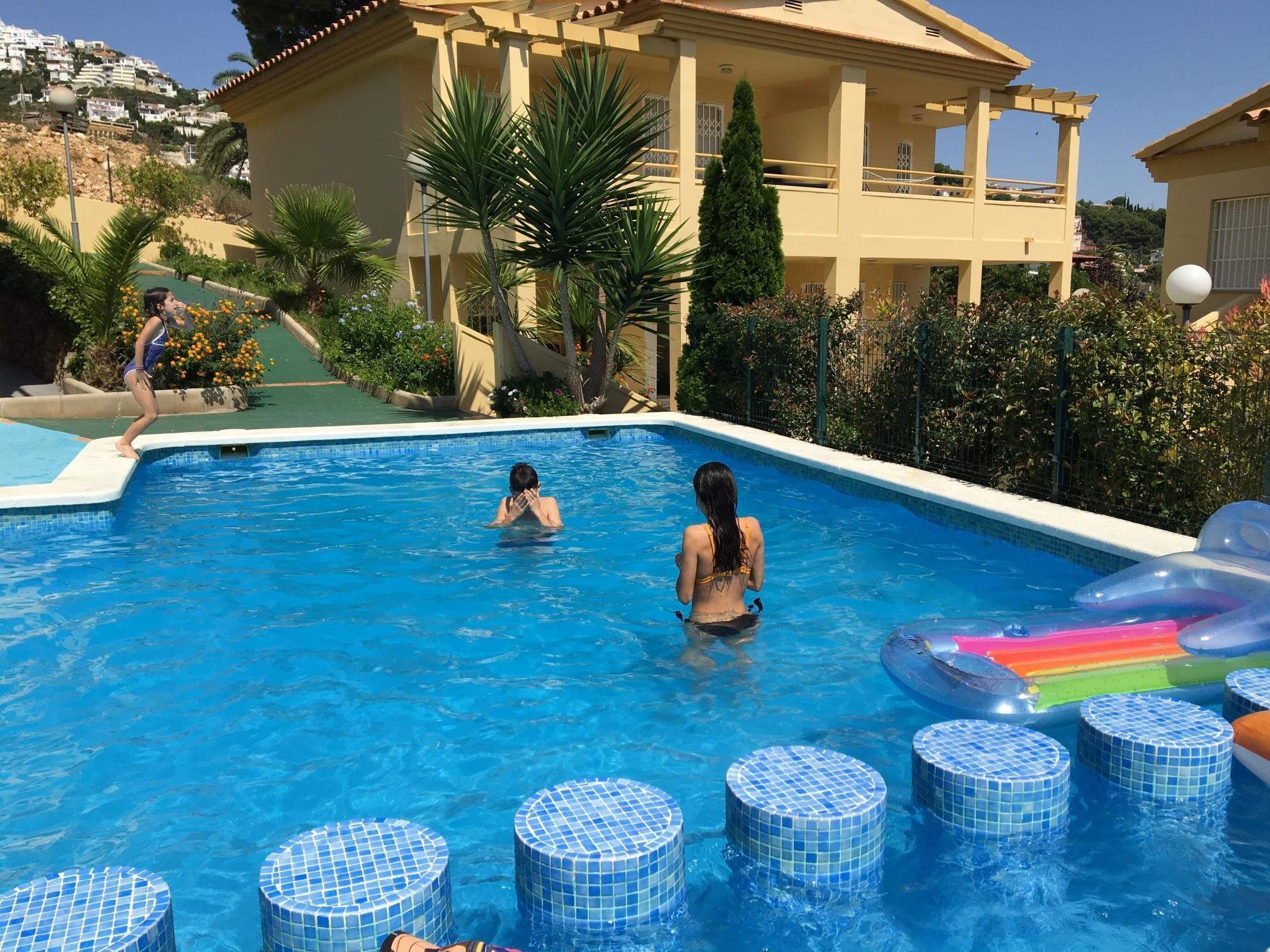 Una piscina con escalones de cerámica que bajan al agua, rodeada de follaje y un edificio con balcón.