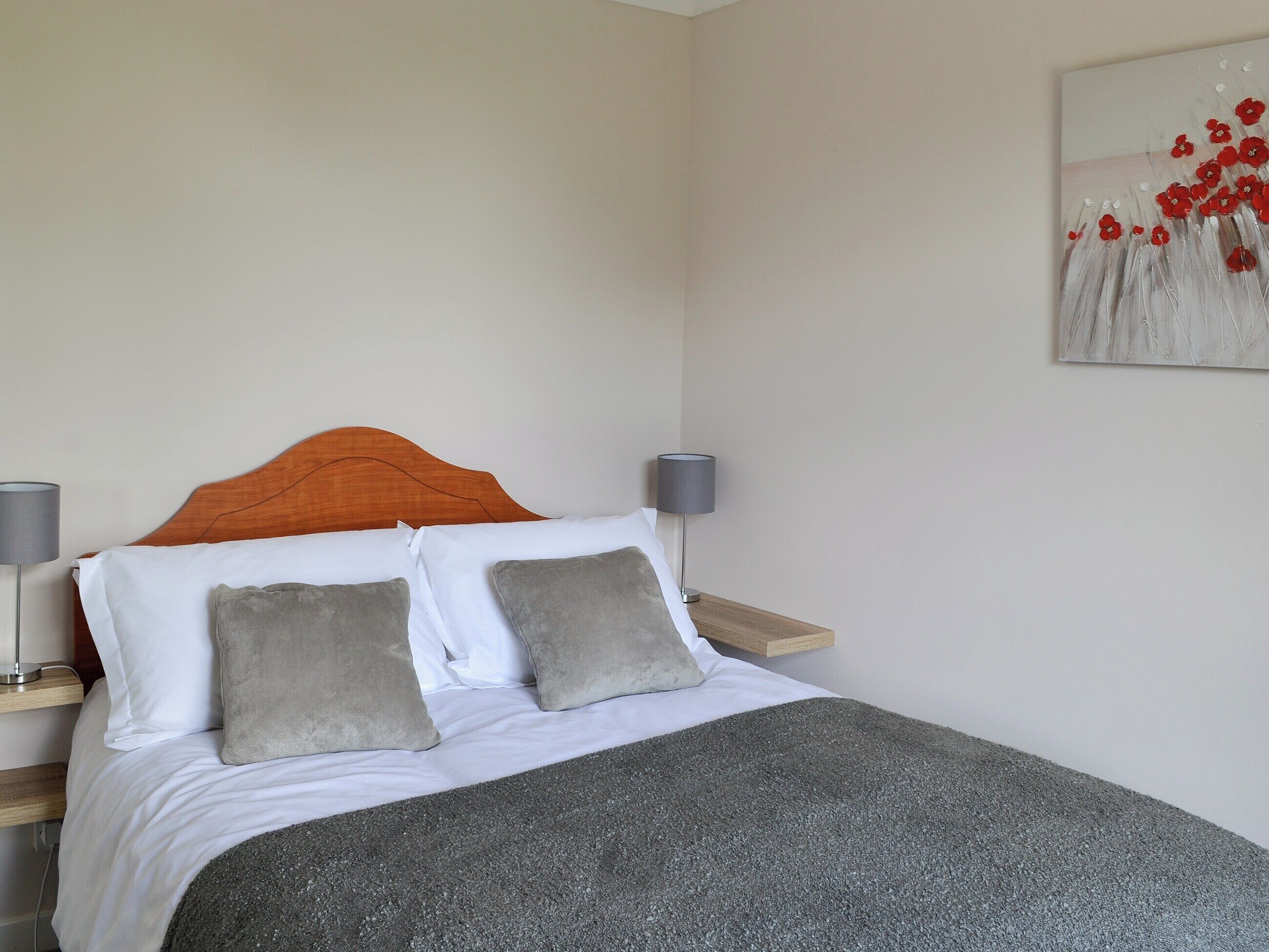 A neatly made bed with a grey blanket, two grey pillows, and an orange headboard. There are two bedside tables with lamps, and a painting of red flowers on the wall.