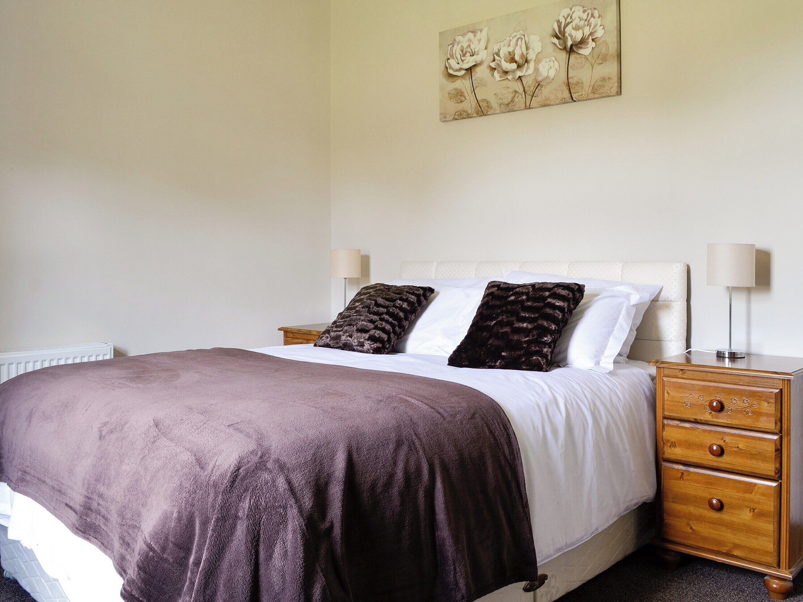 A neatly made bed with a brown blanket, two pillows, a wooden nightstand with a lamp, and a wall-mounted painting of flowers.