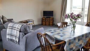 A living room with a sofa, a table with a quilted tablecloth, a wooden chair, a television on a stand, and a vase with flowers. - Little Barn Tynely Farm (Tynely, near Embleton)