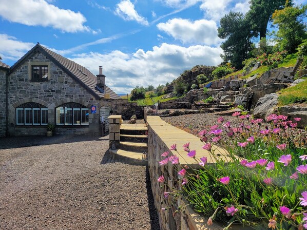 A stone building with a sloped roof and a chimney, a gravel driveway, and a stone wall with pink flowers. - 3 bedroom accommodation in Rothbury (Rothbury)