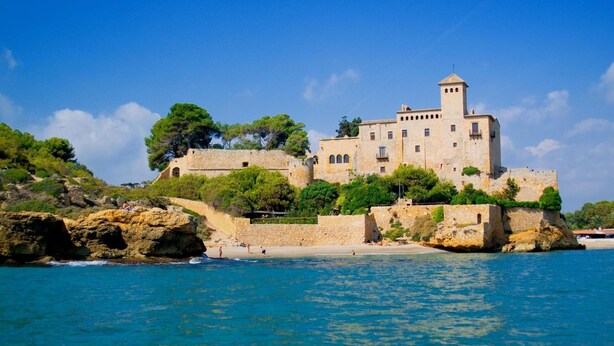 Un castillo de piedra histórico con múltiples torres, situado en una costa rocosa con aguas azul cristalino.