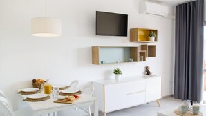 Salón moderno y minimalista con una mesa de comedor blanca, sillas blancas, un mueble blanco, un televisor montado en la pared y una mesita auxiliar con una planta y una taza.