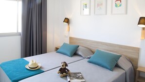 Een netjes opgemaakt bed met een lichtblauwe sprei, twee bijpassende kussens, een klein houten nachtkastje met een lamp en een muur met drie ingelijste foto's.