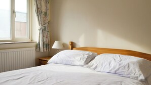 Een opgemaakt bed met witte lakens in een kamer, met een houten hoofdbord, een bedlampje en een raam met gordijnen.