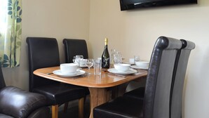 Un coin repas avec une table en bois dressée pour quatre personnes, des chaises en cuir noir, une bouteille de champagne et un téléviseur à écran plat fixé au mur.