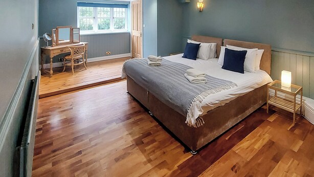 Een slaapkamer met een groot bed, houten vloer, een bureau met spiegel en een raam met uitzicht op groen.